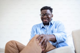 Foto de un hombre africano sentado en un sofá en la sala de estar de su casa y tocándose la rodilla por el dolor durante el día. hombre masajeando su rodilla dolorida.