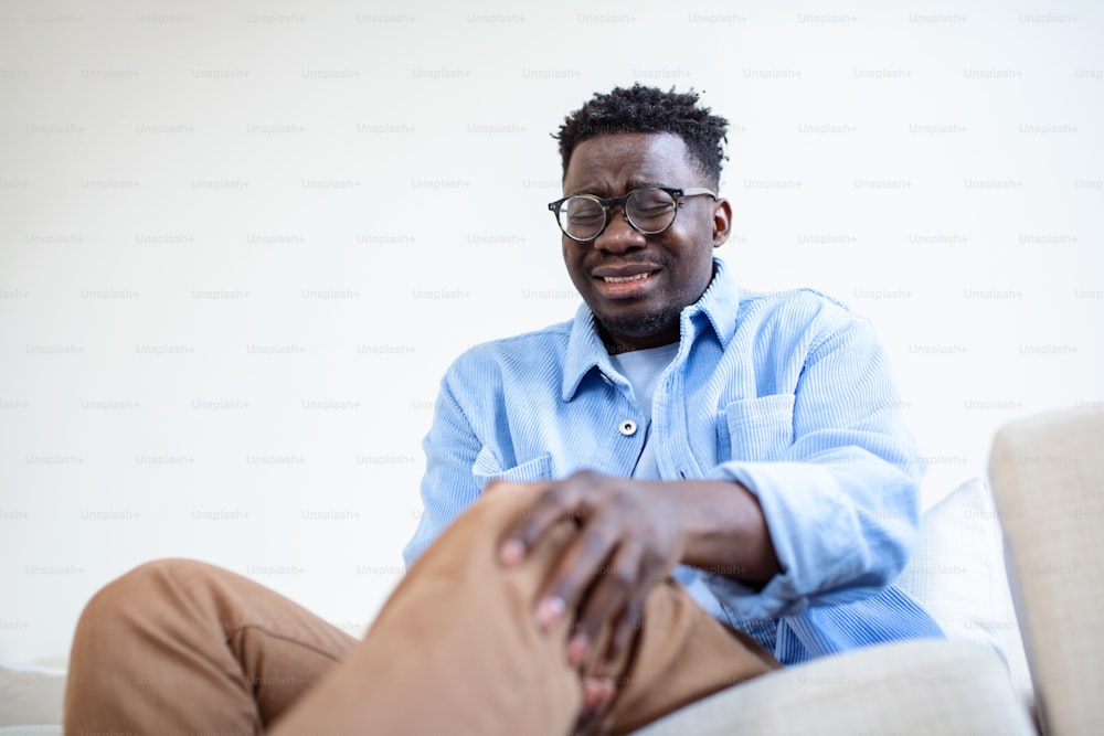 自宅のリビングのソファに座り、日中の痛みで膝を触っているアフリカ人男性の写真。痛む膝をマッサージする男。