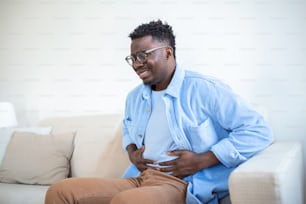 Giovane uomo africano che tiene lo stomaco nel dolore, Uomo con la malattia da reflusso che tiene la pancia con l'espressione dolorosa