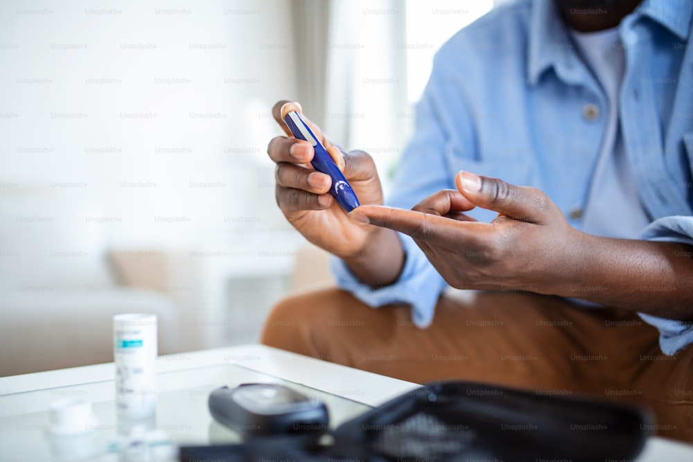 Un hombre africano está sentado en el sofá de la casa y se saca sangre de un dedo debido a la diabetes. La vida cotidiana de un hombre de etnia afroamericana con una enfermedad crónica que está usando un medidor de glucosa.