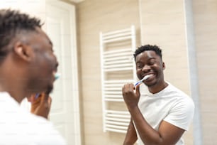 歯ブラシで歯を磨き、バスルームで鏡を見ている笑顔のアフリカ人男性。朝、浴室で歯を磨く男性。夜寝る前に歯を磨くパジャマの男。