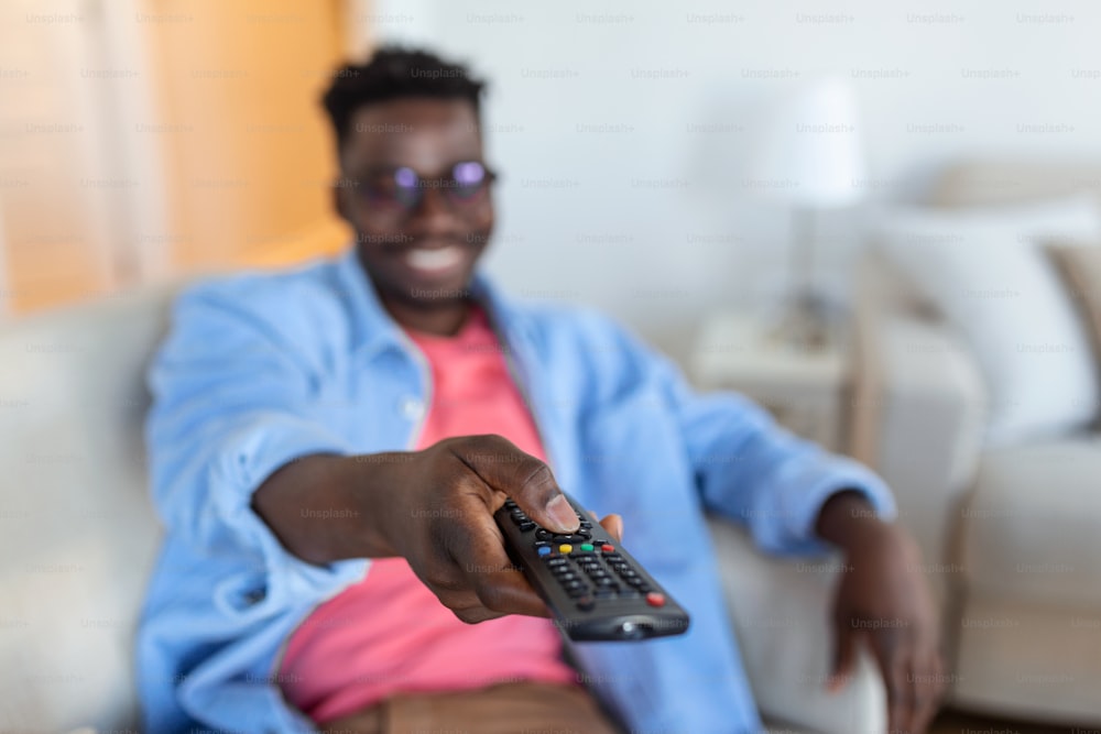 興奮したアフリカ系アメリカ人の男性は、テレビを見ているチャンネルを切り替えるカメラにテレビコントローラーを向けています。テレビ番組の広告。セレクティブフォーカス
