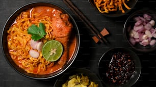 Khao Soi Kai, cuisine unique du nord de la Thaïlande servie avec un plat d’accompagnement sur fond noir. Vue de dessus