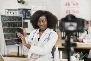 Operatrice medica africana che mostra la scansione a raggi X durante la registrazione di video su una telecamera moderna. Medico qualificato in camice da laboratorio che parla di varie diagnosi.