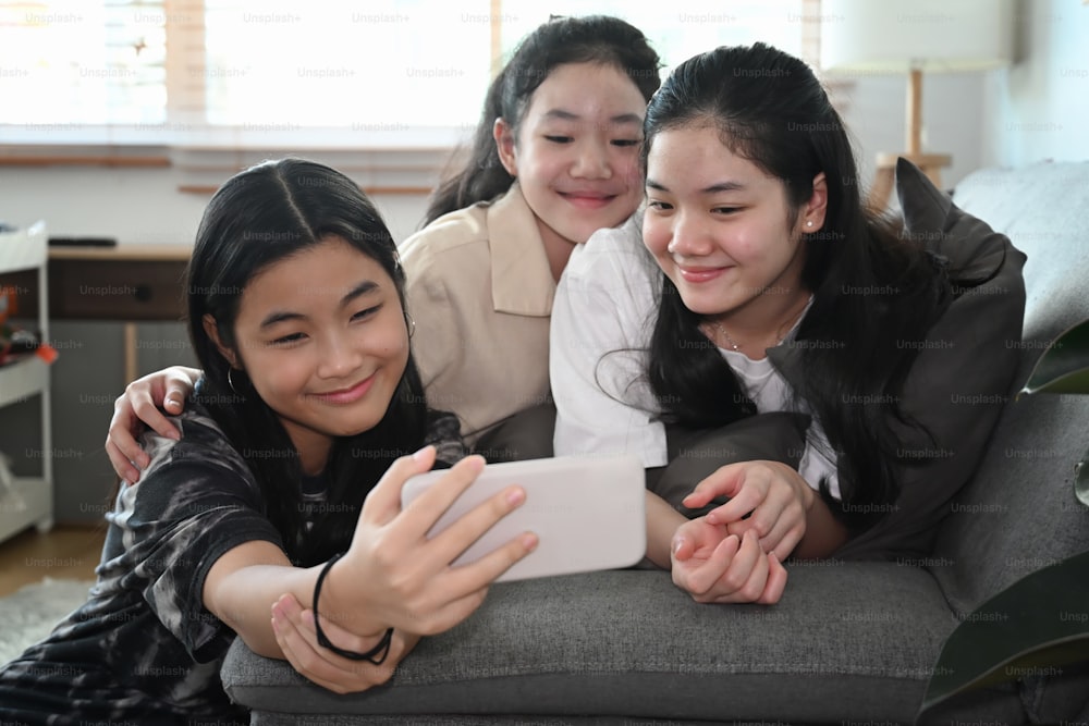 Cheerful Asian girls taking selfie with smart smartphone while sitting together in living room.