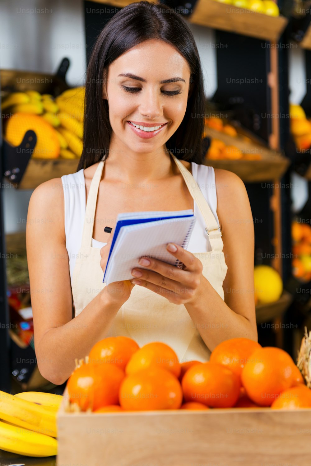 Schöne junge Frau in Schürze, die Notizen im Notizblock macht und lächelt, während sie im Supermarkt mit verschiedenen Früchten im Hintergrund steht