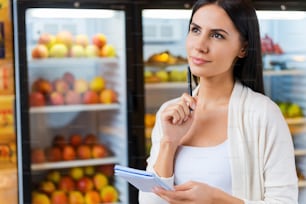 ��買い物リストを持ち、食料品店の冷蔵庫の前に立ちながら目をそらす思慮深い若い女性