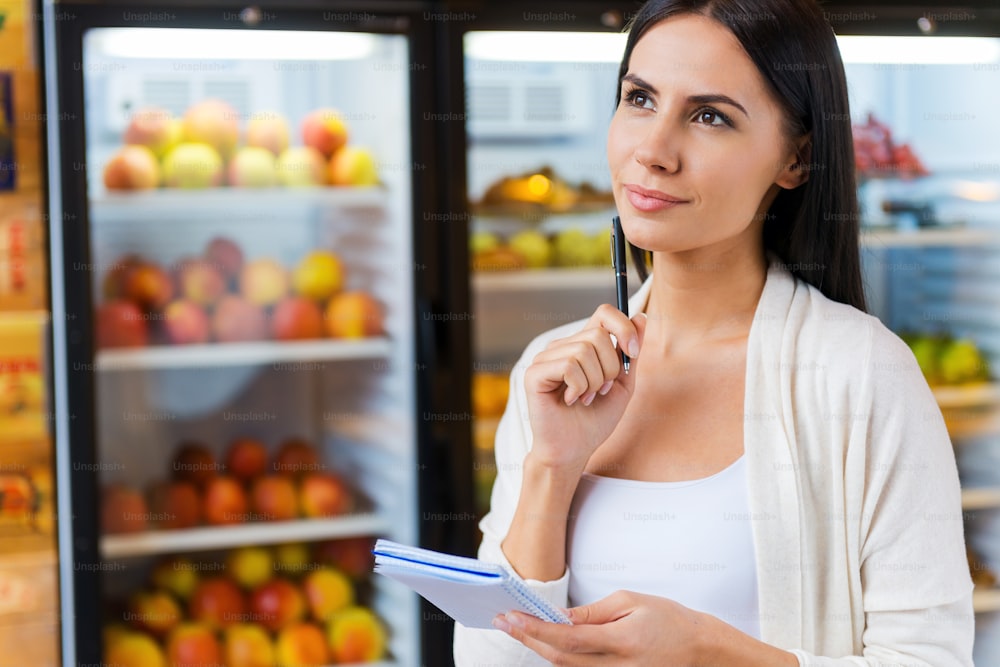 Thoughtful young woman holding shopping list and looking away while standing in front of refrigerators in grocery store