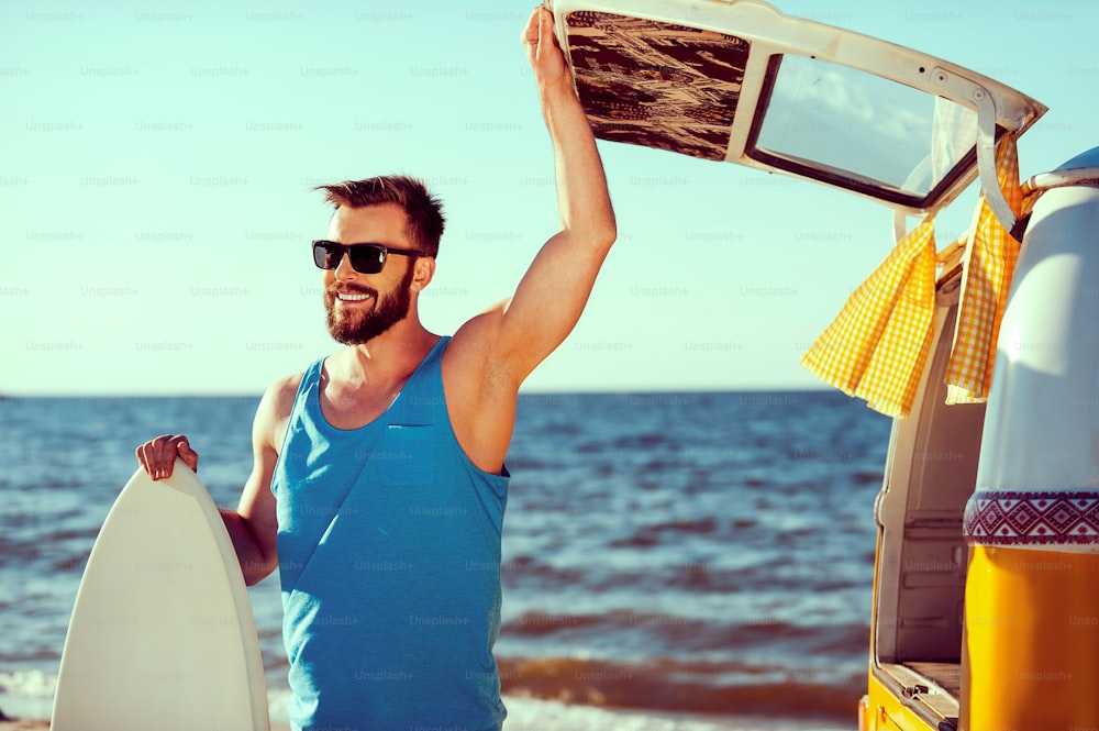 Smiling young man holding skimboard and while opening a trunk door of his retro minivan with sea in the background