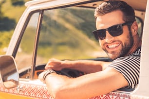 Cheerful young man smiling at camera and holding hand on steering wheel while sitting inside of his minivan