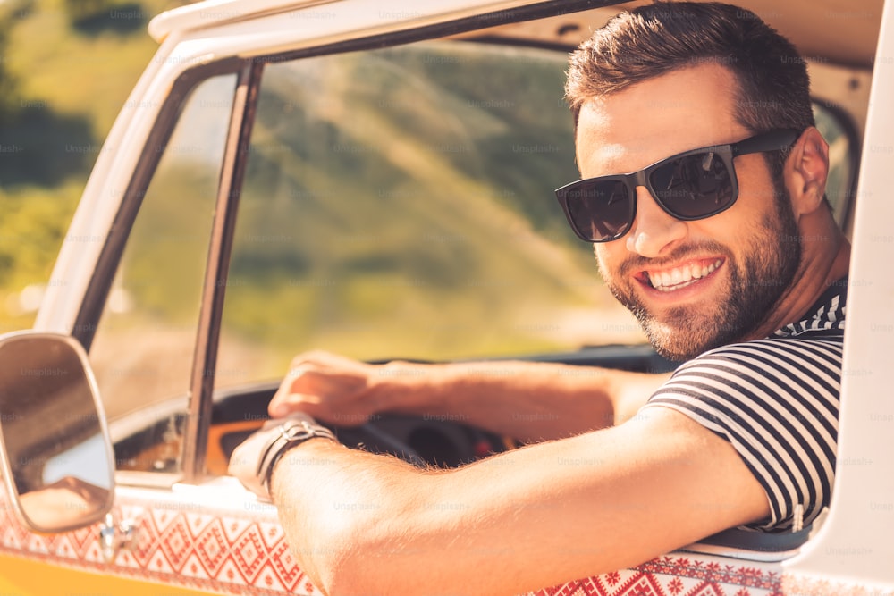 Cheerful young man smiling at camera and holding hand on steering wheel while sitting inside of his minivan