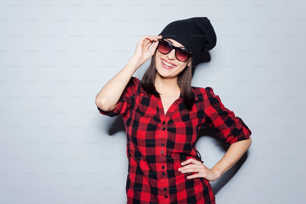 Retrato de una hermosa mujer joven ajustándose sus gafas y sonriendo mientras está de pie sobre fondo gris