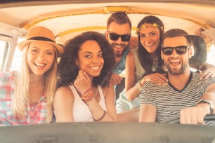 Grupo de jovens alegres sorrindo para a câmera enquanto estão sentados dentro da minivan juntos