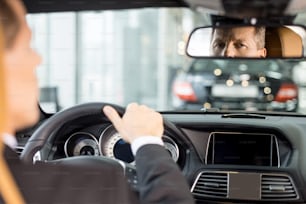 Vista traseira do homem sênior confiante em formalwear sentado no lugar dos motoristas no carro e olhando para o espelho