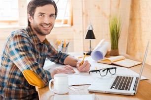 Seitenansicht eines lächelnden jungen Mannes, der zeichnet und in die Kamera schaut, während er an seinem Arbeitsplatz sitzt