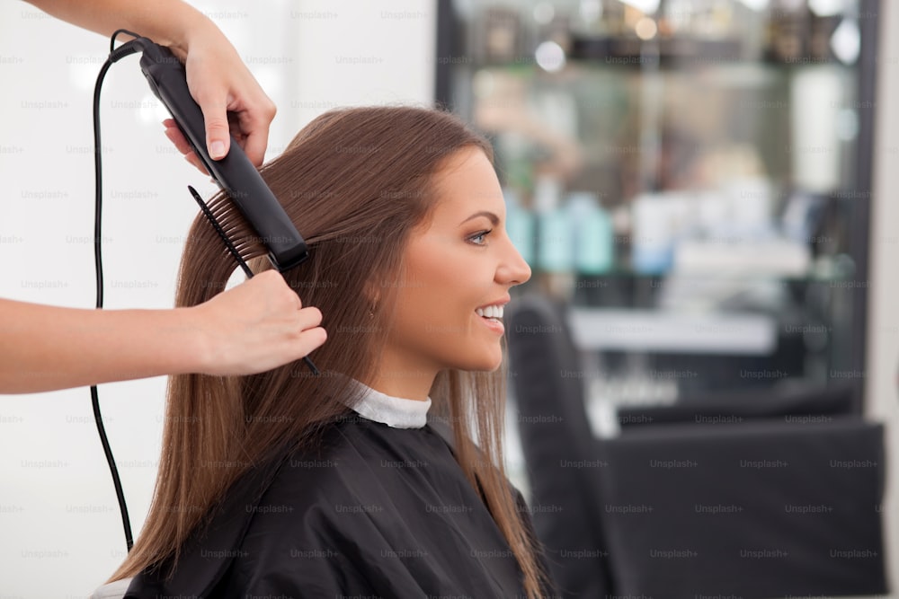 Pretty young woman is getting her hair equalized in beauty salon. The hairdresser is combing her hair and holding an iron. The female client is smiling. Copy space in right side