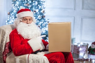 Old Santa Claus is sitting in a chair and smiling. He is holding a box of gif and pointing his finger at it with joy. There is a holiday tree on the background