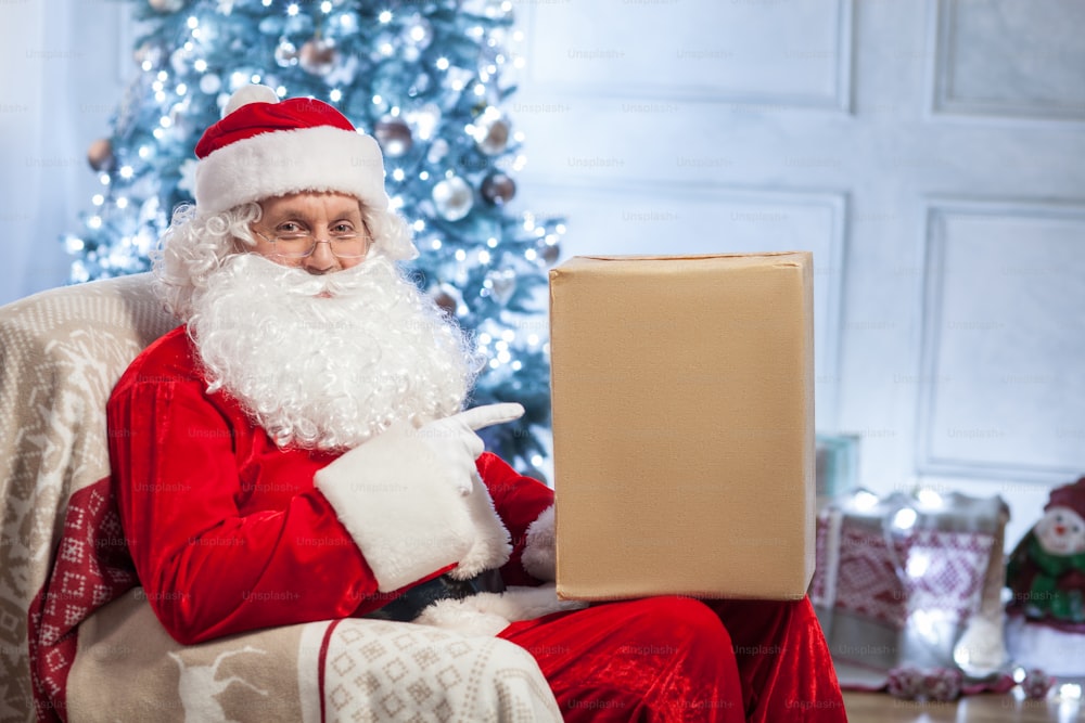 Old Santa Claus is sitting in a chair and smiling. He is holding a box of gif and pointing his finger at it with joy. There is a holiday tree on the background