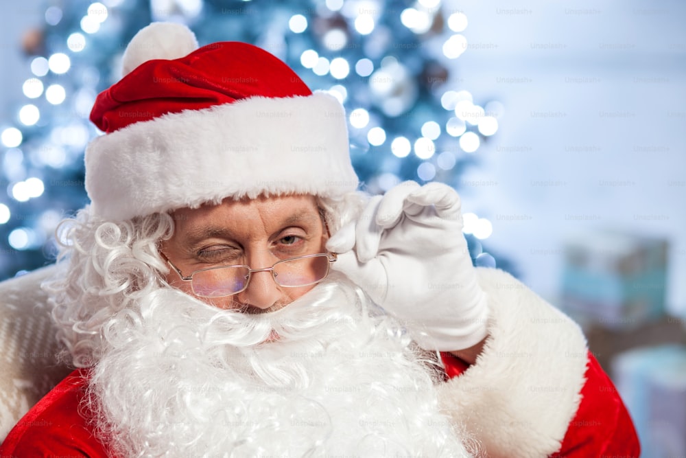 Generous Santa Claus is looking forward and winking. He is touching his eyeglasses and smiling. The old man is sitting in his chair near a fir-tree and presents
