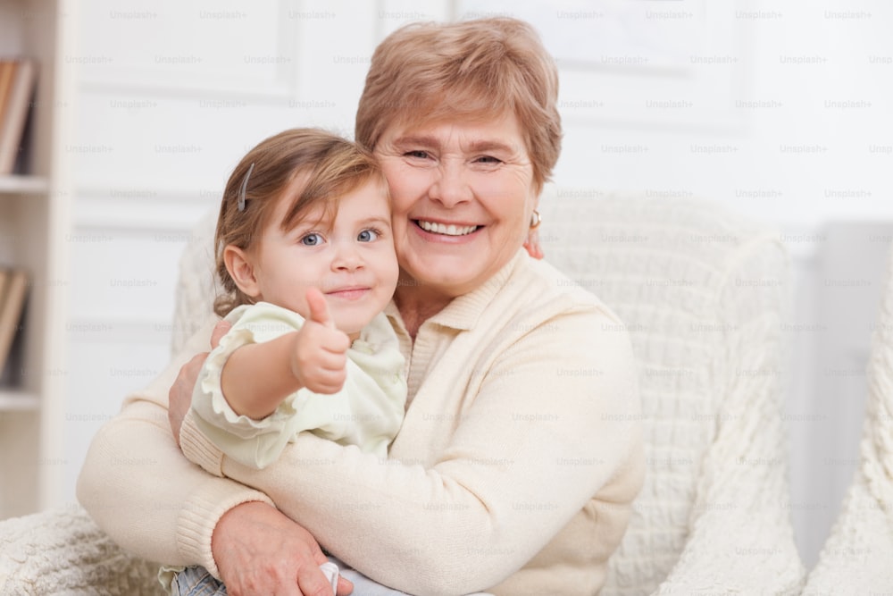 Cheerful grandmother is caring of her granddaughter. They are sitting on the sofa and embracing. The girl is giving thumb up. They are smiling