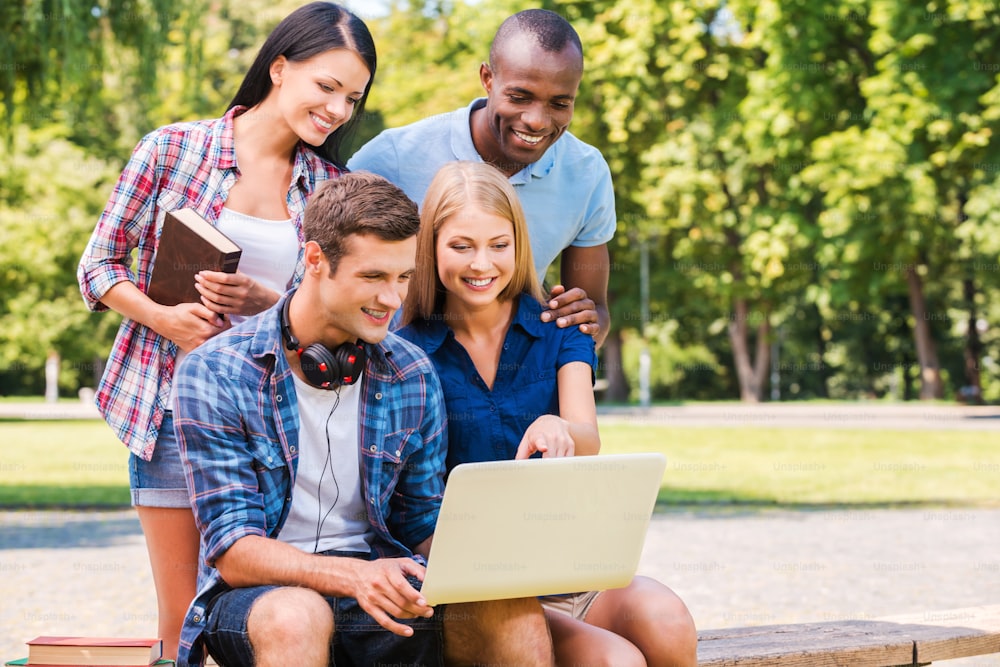 Vier fröhliche junge Leute, die etwas diskutieren und auf den Laptop schauen, während sie zusammen draußen sitzen