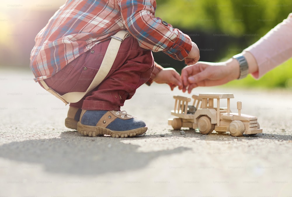 A close-up of an adult helping a young boy play with a wooden tractor on pavement in an outdoor setting.  The boy is visible from the waist down, and only the adult's right arm is shown.
