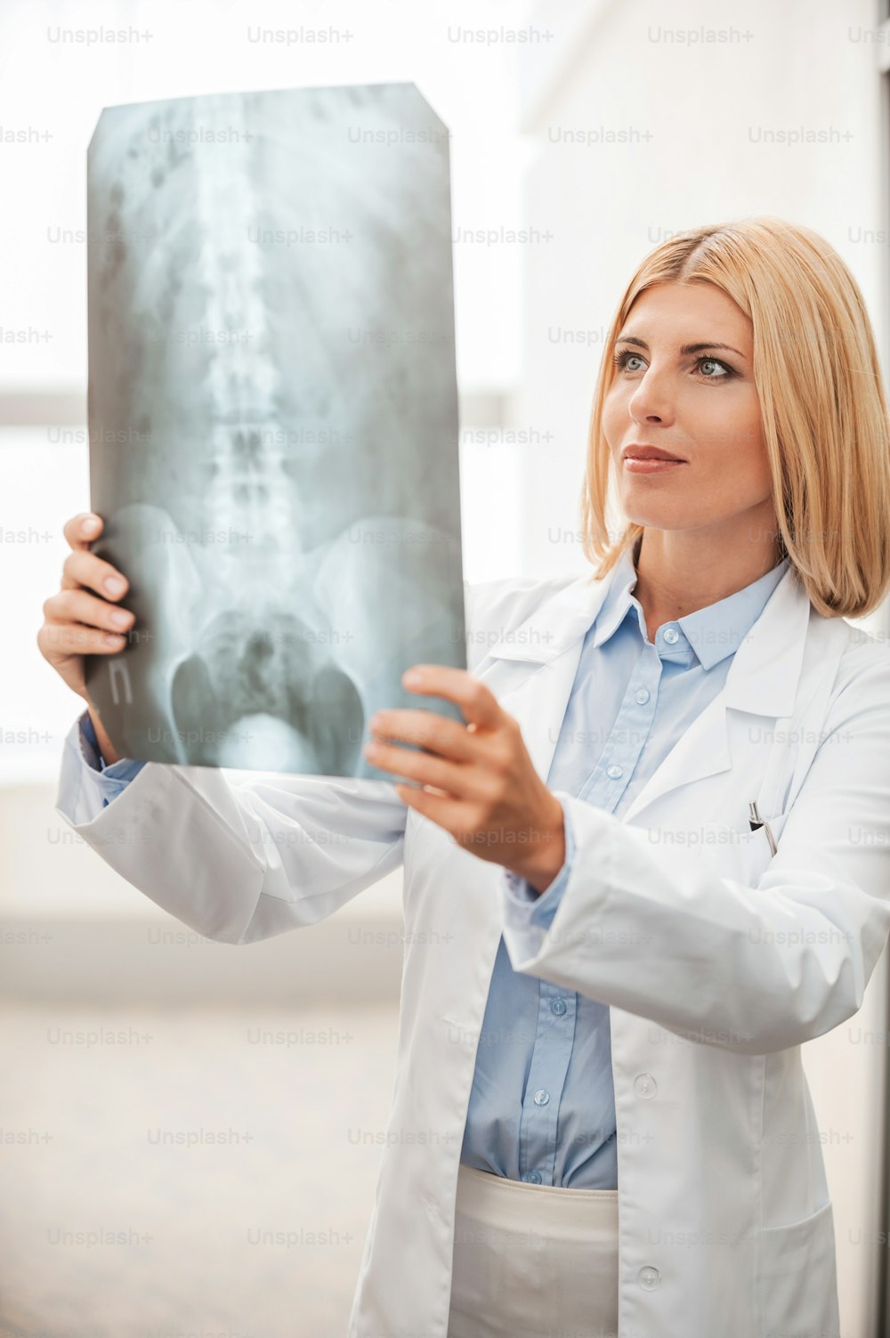 Médico examinando radiografías. Doctora segura de sí misma en uniforme blanco sosteniendo una imagen de rayos X y mirándola