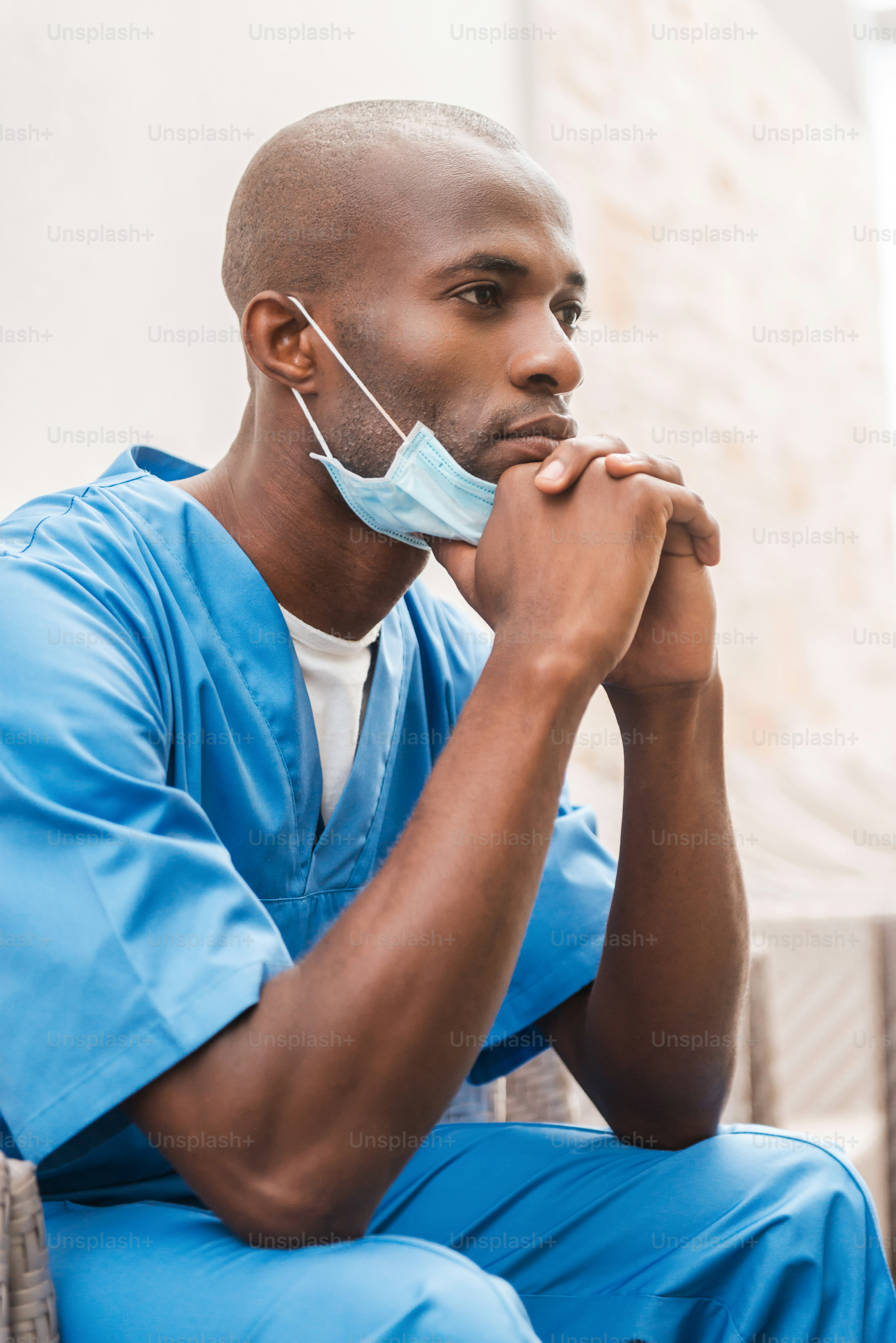 Thoughtful surgeon. Thoughtful young African doctor in blue uniform leaning his face on hands and looking away while sitting at the chair in corridor
