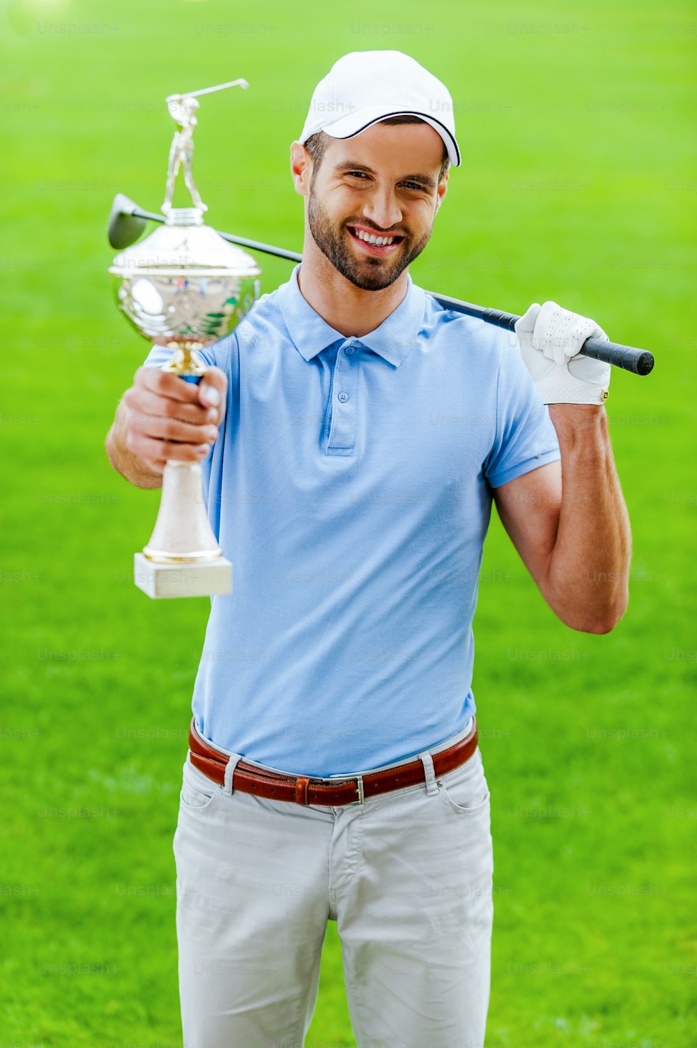 Golfista masculino feliz mostrando su trofeo y llevando al conductor al hombro mientras está de pie en el campo de golf