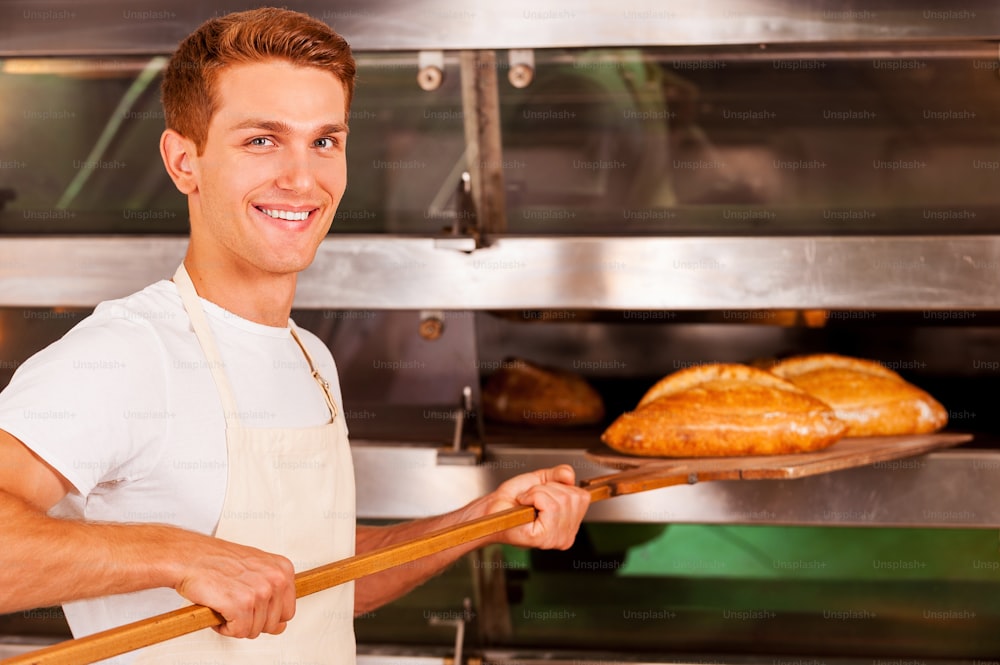 Confident young man in apron taking fresh baked bread from oven and smiling