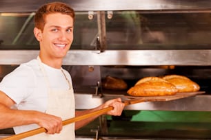 Selbstbewusster junger Mann in Schürze nimmt frisch gebackenes Brot aus dem Ofen und lächelt