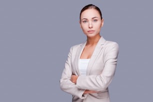 Confident young businesswoman keeping arms crossed while standing against grey background