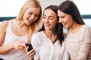 Drei schöne junge Frauen, die aufs Handy schauen und lächeln, während sie zusammen auf der Couch sitzen