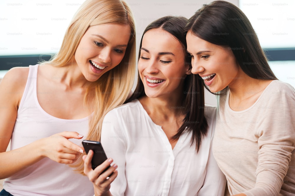 Three beautiful young women looking at the mobile phone and smiling while sitting on the couch together