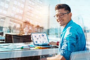 Bon travailleur. Beau contenu jeune homme souriant et portant des lunettes tout en travaillant sur son ordinateur portable et ses affaires posées sur la table