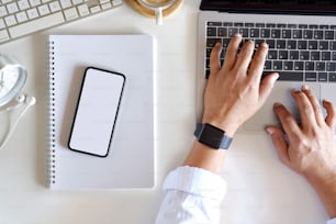 Cropped shot of man working by using a laptop computer on white table. Hands typing on a keyboard and office supplies. Top view.