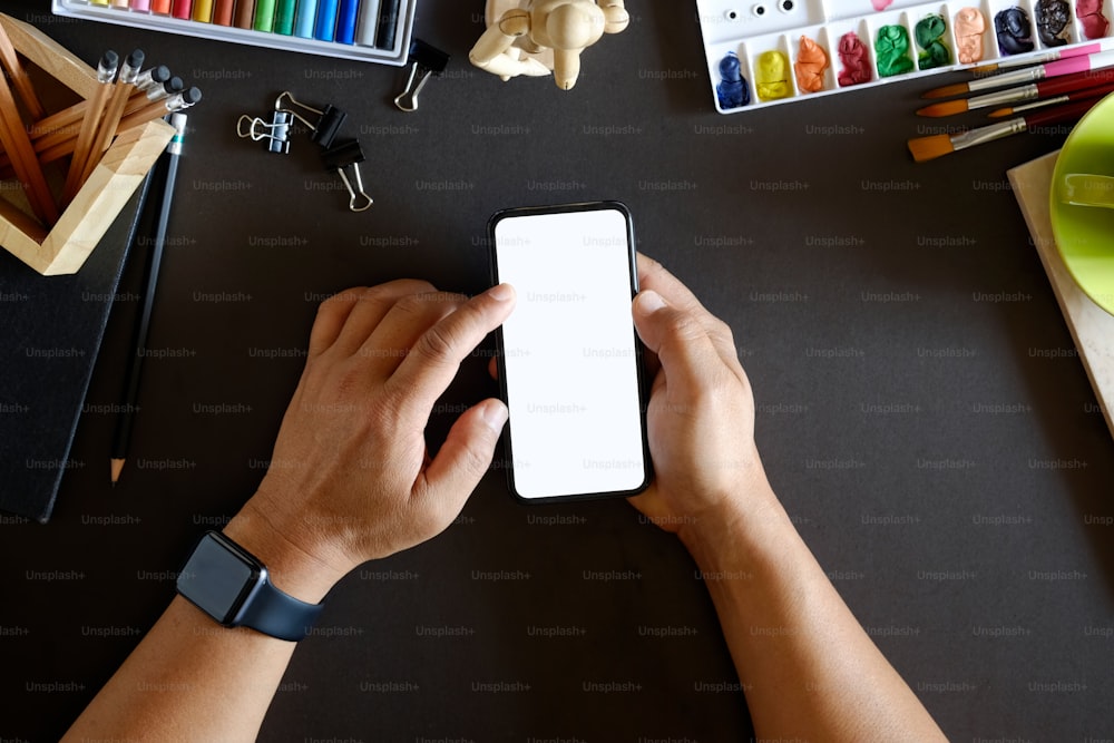 暗いデザイナーの机、ラップトップ、アーティストの工芸品、携帯電話に空白のモックアップ画面がある上でスマートフォンを使用している男性のトリミングされたトップビューショット。