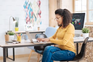 Wonderful day. Pretty joyful content young curly-haired woman smiling and working on her laptop while sitting at the table