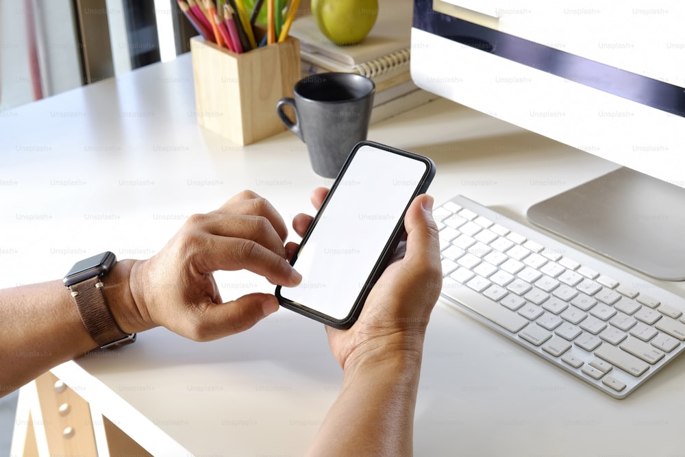 Foto recortada de un hombre sosteniendo un teléfono móvil con pantalla en blanco sobre el escritorio. Teléfono inteligente con pantalla para montaje de visualización gráfica.