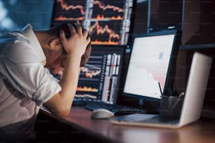 Stressful day at the office. Young businessman holding hands on his face while sitting at the desk in creative office. Stock Exchange Trading Forex Finance Graphic Concept.