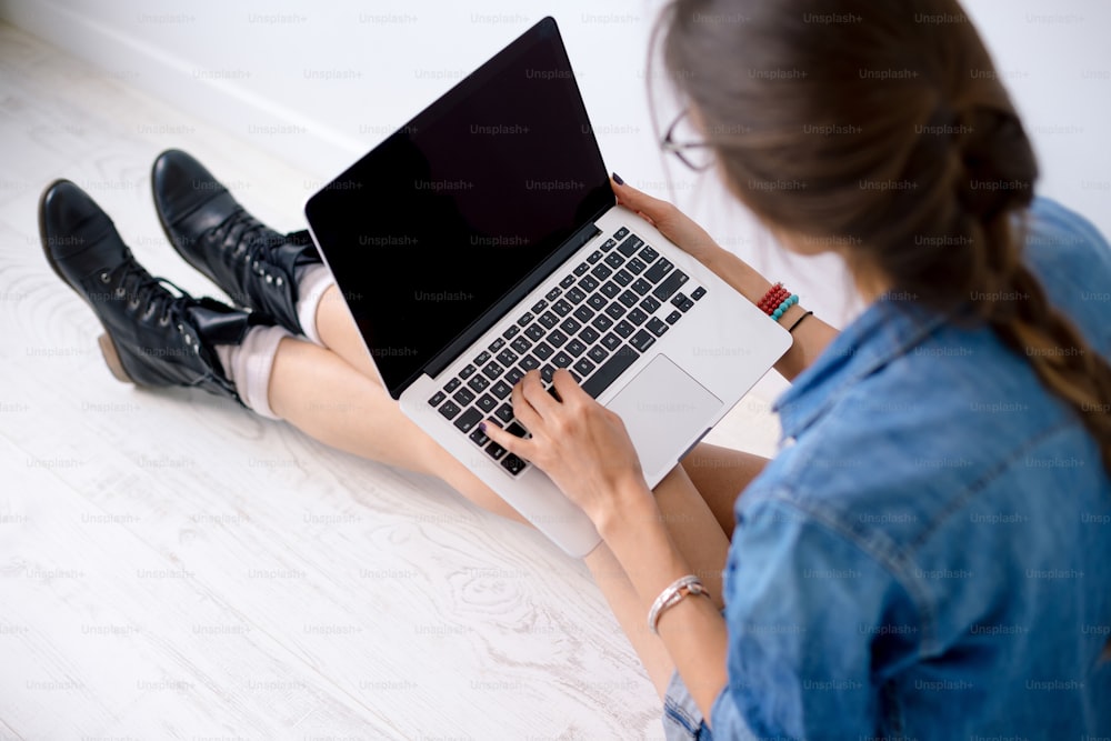 Close-up portrait of womans hands working on her laptop keyboard, while sitting on white flor holding the computer on her long legs.