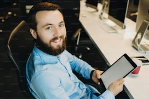 Homme joyeux non rasé en chemise bleue assis confortablement sur une chaise de bureau sur le lieu de travail tenant dans les mains une tablette et souriant en regardant la caméra
