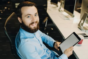 Cheerful unshaven male in blue shirt sitting comfortably in office chair at workplace holding in hands tablet and smiling looking at camera