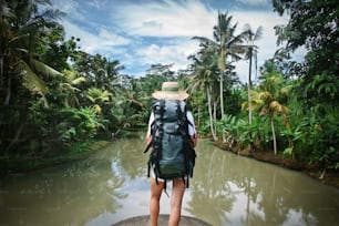 Woman traveler with backpack and hat standing near tropical river