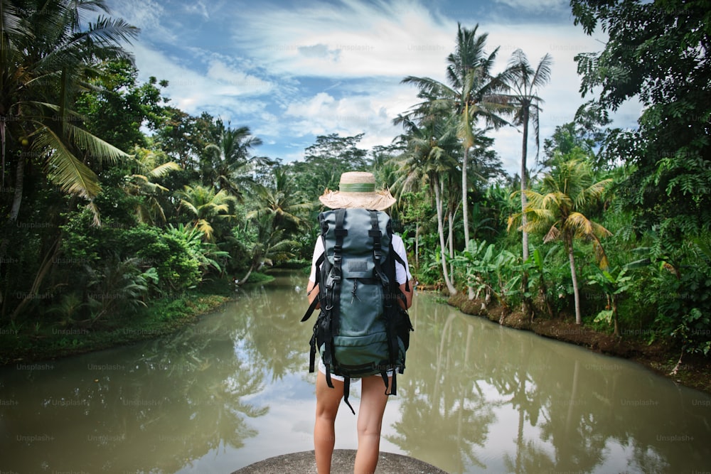 Woman traveler with backpack and hat standing near tropical river