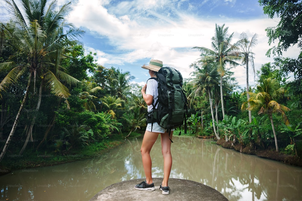 Mulher viajante corajosa com mochila em pé na borda perto do grande rio tropical