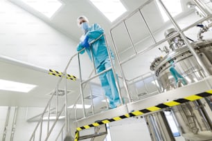 cientista está de pé na escada, olhe para a câmera, traje de laboratório azul, mãos cruzadas, perto de tanques cromados