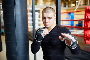 戦いのポーズに立って、スポーツクラブでボクシングの練習中にカメラを見ている決意の若い筋肉の男の肖像画