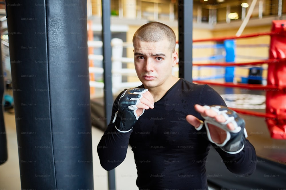 Portrait of determined young muscular man standing in fight pose and looking at camera  during boxing practice in sports club
