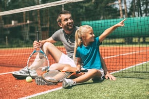 쾌활한 아버지와 딸은 테니스 네트에 기대어 테니스 코트에 앉아 미소를 지으며 멀리 바라보고 있습니다.